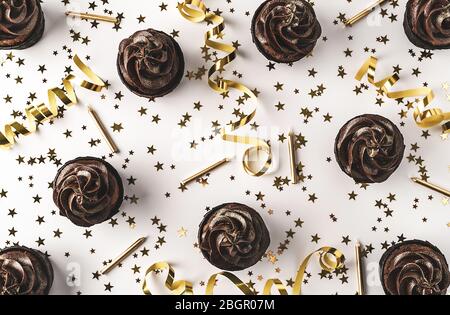 Cupcake al cioccolato con glassa al cioccolato e spruzzati d'oro su sfondo bianco decorati con confetti a forma di stella dorata, candele e festa Foto Stock