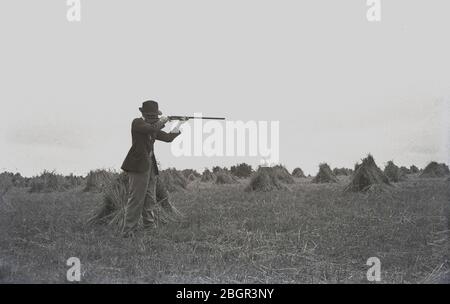 Anni '1930, storico agricoltore gentiluomo che indossa una giacca sportiva e un cappello in piedi in un campo di fieno falciato con piccoli fieno, con il suo fucile tenuto in alto mirato a un bersaglio, forse un coniglio, Devon, Inghilterra, Regno Unito. Foto Stock