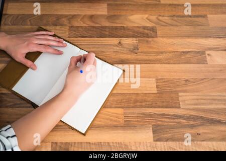 Vista superiore di una donna che scrive in un notebook su un tavolo di legno nella sua cucina domestica. Lavoro a casa e soggiorno a casa. Foto Stock