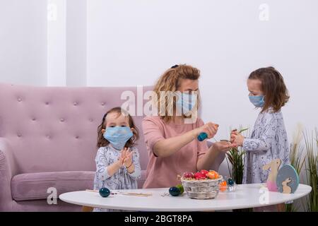 Madre e figlie stanno dipingendo le uova. Famiglia felice si prepara per Pasqua. Madre che indossa la maschera di sua figlia contro il coronavir. Quarantena a hom Foto Stock