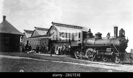 Negli anni '10, una locomotiva a vapore, con la sua vettura a carbone e una singola vettura per passeggeri in un piccolo deposito ferroviario cittadino nel Wisconsin settentrionale, America. Non c'è caboose collegato, perché questo sarebbe stato un piccolo treno utilizzato per brevi tratti tra molte città e villaggi del nord. L'edificio del deposito della stazione, sulla sinistra, è ingenuito da tre grandi fienili lungo i binari. Loro furono probabilmente usati per immagazzinare mais o prodotti di fattoria che necessitavano di essiccazione. Quindici passeggeri sono in attesa, mentre un ingegnere si trova accanto al motore. Per vedere le mie immagini relative al trasporto, Cerca: Prestor veicolo d'epoca Foto Stock