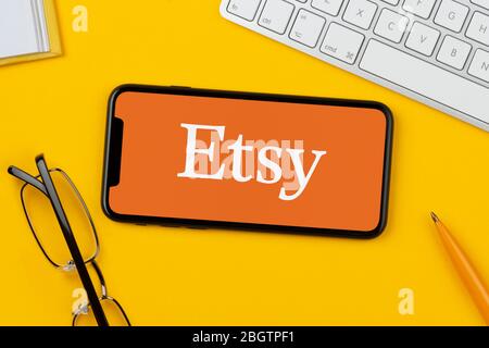 Uno smartphone con il logo Etsy è posizionato su uno sfondo giallo insieme a tastiera, occhiali, penna e libro (solo per uso editoriale). Foto Stock