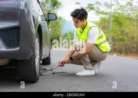 Giovane uomo asiatico con giubbotto di sicurezza verde che sostituisce il pneumatico forato sulla sua auto allentando i dadi con una chiave per ruota prima di sollevare il veicolo Foto Stock