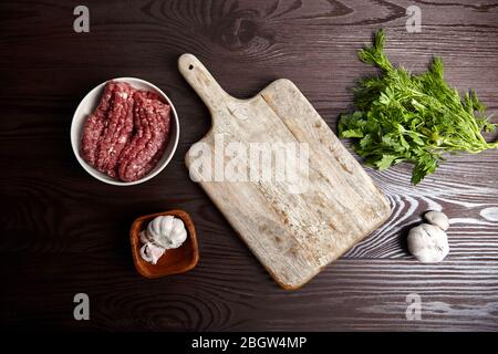 Cottura di carne macinata. Tagliere con condimenti freschi su un tavolo di legno, piatto. Teste e spicchi d'aglio bianchi, foglie di aneto verde e prezzemolo. Bo Foto Stock