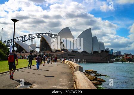 Il turismo a Sydney con i visitatori che camminano lungo il lungomare del porto di Sydney con vista dell'iconica Sydney Opera House e del Ponte dell'Porto. Foto Stock
