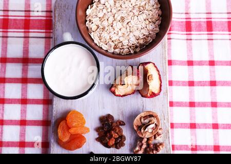 Ciotola di farinata d'avena, tazza di yogurt, marmellata, cioccolato, uvetta, albicocche secche e noci su tagliere di legno su sfondo tessuto a scacchi Foto Stock