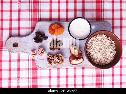 Ciotola di farinata d'avena, tazza di yogurt, marmellata, cioccolato, uvetta, albicocche secche e noci su tagliere di legno su sfondo tessuto a scacchi Foto Stock