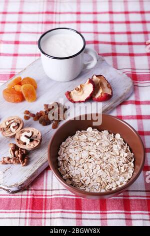 Ciotola di farinata d'avena, tazza di yogurt, marmellata, cioccolato, uvetta, albicocche secche e noci su tagliere di legno su sfondo tessuto a scacchi Foto Stock