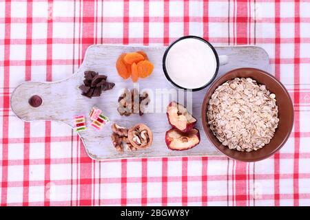 Ciotola di farinata d'avena, tazza di yogurt, marmellata, cioccolato, uvetta, albicocche secche e noci su tagliere di legno su sfondo tessuto a scacchi Foto Stock