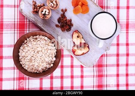 Ciotola di farinata d'avena, tazza di yogurt, marmellata, cioccolato, uvetta, albicocche secche e noci su tagliere di legno su sfondo tessuto a scacchi Foto Stock