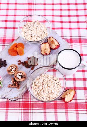 Ciotola di farinata d'avena, tazza di yogurt, marmellata, cioccolato, uvetta, albicocche secche e noci su tagliere di legno su sfondo tessuto a scacchi Foto Stock