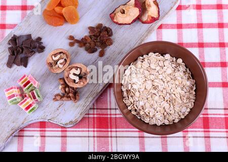 Ciotola di farinata d'avena, tazza di yogurt, marmellata, cioccolato, uvetta, albicocche secche e noci su tagliere di legno su sfondo tessuto a scacchi Foto Stock