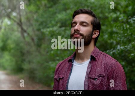 giovane uomo con capelli e barba marroni che indossa il pulsante rosso in basso respira in un soffio di aria fresca immergersi in un percorso naturale a distanza sociale hi Foto Stock