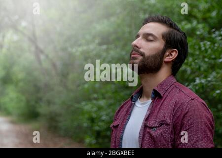 giovane uomo con capelli e barba marroni che indossa il pulsante rosso in basso respira in un soffio di aria fresca immergersi in un percorso naturale a distanza sociale hi Foto Stock