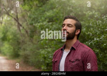 giovane uomo con capelli e barba marroni che indossa il pulsante rosso in basso respira in un soffio di aria fresca immergersi in un percorso naturale a distanza sociale hi Foto Stock