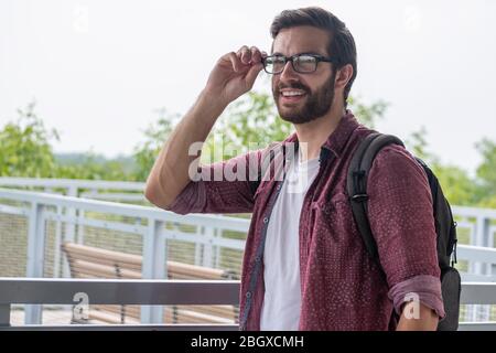 Un hipster bearded che indossa una camicia rossa del tasto giù che porta i suoi occhiali prescritti per vedere più chiaramente che cosa è nella distanza con una stare diritta Foto Stock