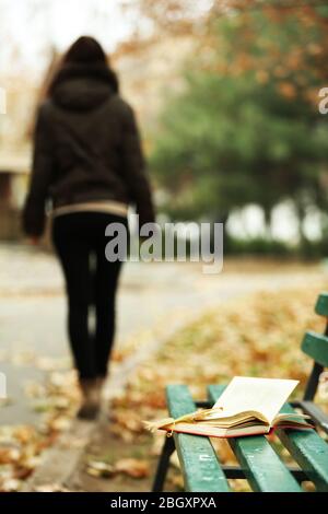 Libro a sinistra sulla panchina con silhouette di ragazza a piedi nel parco autunnale Foto Stock