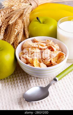 Fiocchi di mais dolci in ciotola ceramica, frutta e bicchiere con latte su tovagliolo, su sfondo legno colorato Foto Stock