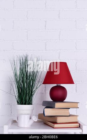 Interni con lampada, pianta, vaso di irrigazione in ceramica e libri su piano tavolo su sfondo bianco muro di mattoni Foto Stock