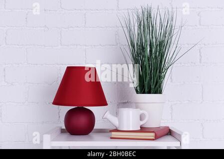 Interni con lampada, pianta, vaso di irrigazione in ceramica e libro su piano tavolo su sfondo bianco muro di mattoni Foto Stock