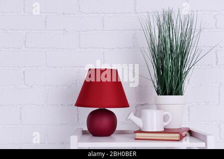 Interni con lampada, pianta, vaso di irrigazione in ceramica e libro su piano tavolo su sfondo bianco muro di mattoni Foto Stock