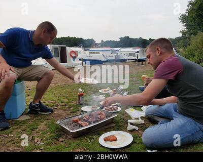 Due ragazzi che godono di un barbecue sulle rive del Norfolk Broads mentre ormeggiato in su a Salhouse Broad, Norfolk Broads vacanza in barca Foto Stock