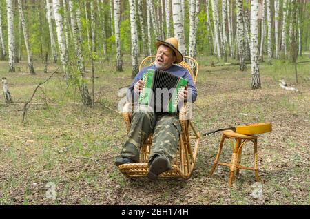 Il caucsiano indossa il cappello di paglia che canta e suona la garmonika mentre siede solitario nella sedia a dondolo di vimini nella foresta di betulla Foto Stock