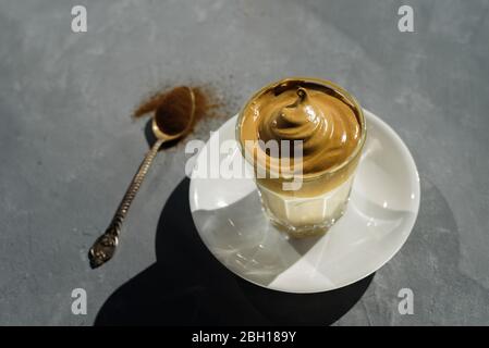 Dalgona caffè istantaneo montato bevanda in vetro. Luce solare dura, fondo in cemento. Con cucchiaino da tè di caffè in polvere Foto Stock