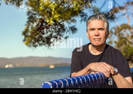 Ritratto di un uomo maturo 57 anni con capelli grigi guardando macchina fotografica seduto su una panchina in una spiaggia all'aperto, sicuro, rilassato Foto Stock