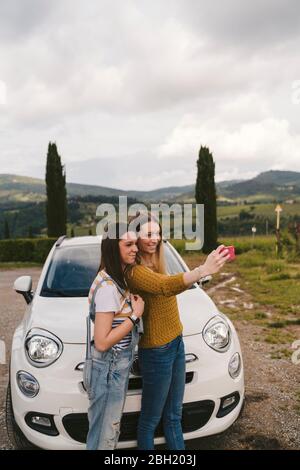 Due felici giovani donne in piedi accanto a auto prendendo un selfie, Toscana, Italia Foto Stock