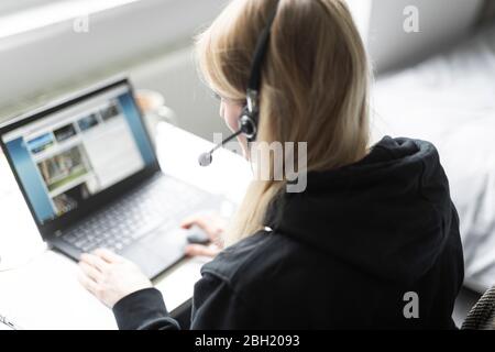 Donna bionda con cuffie sedute alla scrivania a casa che lavorano su un computer portatile Foto Stock