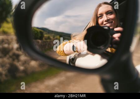 Riflesso nello specchio dell'ala di una giovane donna sorridente con la fotocamera che si appoggia fuori dal finestrino dell'auto Foto Stock