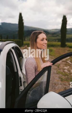 Sorridente giovane donna che esce dall'auto in campagna, Toscana, Italia Foto Stock
