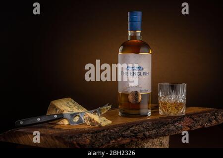 Una bottiglia di whisky inglese e un bicchiere di whisky sul ghiaccio con formaggio blu su un pannello di formaggio di quercia, una combinazione perfetta di whisky inglese e formaggio. Foto Stock