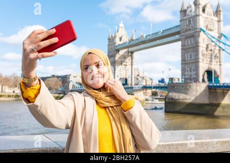Ritratto di giovane donna sorridente che prende selfie con smartphone di fronte a Tower Bridge, Londra, Regno Unito Foto Stock