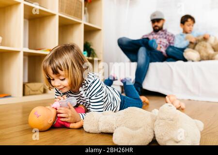 Bambina che la alimenta, mentre fratello e padre sono seduti sul divano Foto Stock