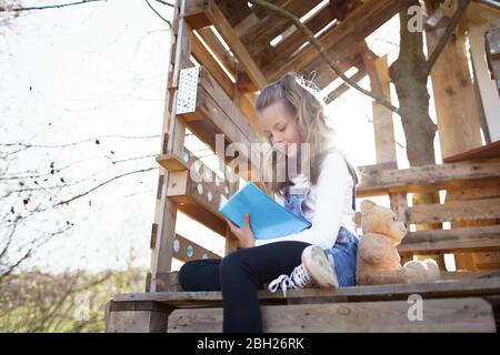 Ragazza seduta in giardino a casa dell'albero e scrivendo qualcosa nel suo diario blu Foto Stock