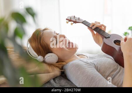 Giovane donna spensierata con cuffie e ukulele a casa Foto Stock