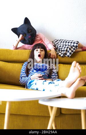 Due ragazze annoiate che si siedono sul divano, guardano la TV, una che indossa una maschera di gatto Foto Stock