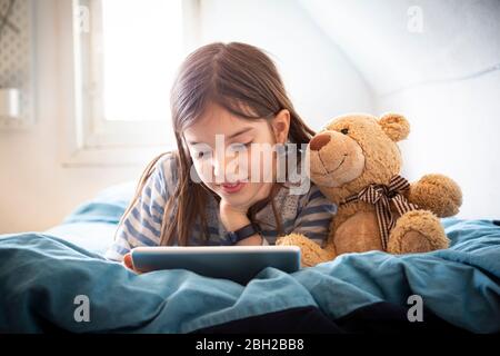 Ritratto di una ragazza sorridente che si stette a letto con orsacchiotto utilizzando un tablet digitale Foto Stock