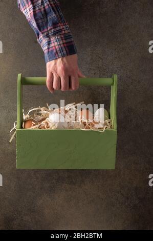 Donna contadina mano tiene una scatola di legno con uova marroni e bianche in paglia su uno sfondo scuro. Orientamento verticale Foto Stock