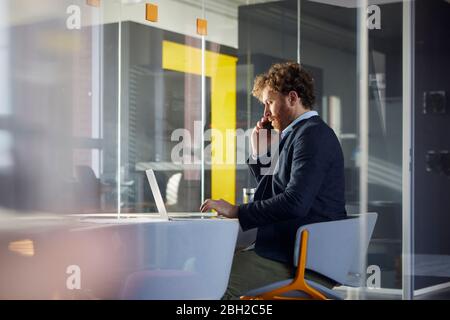 Uomo d'affari seduto alla scrivania in ufficio utilizzando il computer portatile e lo smartphone Foto Stock