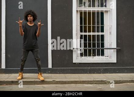 Ritratto di giovane uomo con afro in piedi sul marciapiede che mostra Rock e Roll Sign Foto Stock