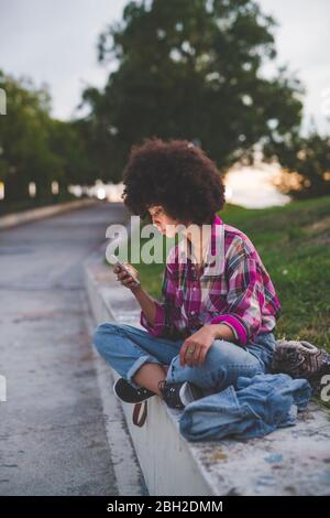 Giovane donna con capelli afro seduta sul marciapiede con smartphone Foto Stock