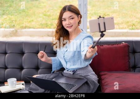 Felice giovane bella donna asiatica che tiene il suo telefono su un selfie bastone per scattare foto o video di se stessa nel suo soggiorno mentre lavora a casa Foto Stock