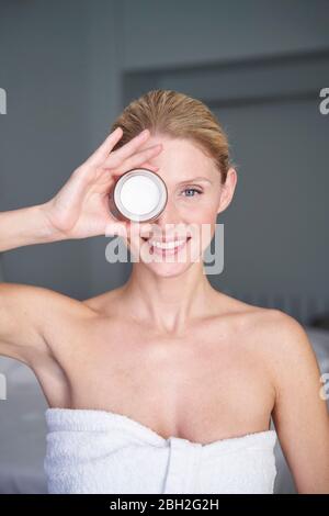 Ritratto di sorridente bella donna che tiene vaso crema davanti al suo occhio Foto Stock