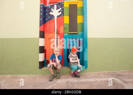 Ritratto di ragazzo e sua sorellina seduti insieme sul gradino di fronte alla colorata porta d'ingresso dipinta Foto Stock