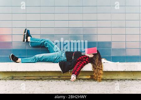 Ragazza adolescente che si trova su un muro all'aperto che copre la faccia con libro Foto Stock