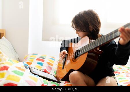 Ragazzo seduto a letto utilizzando un tablet digitale per suonare la canzone sulla chitarra Foto Stock