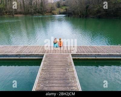 Vista posteriore di due amici seduti fianco a fianco sul molo, Valdemurio Reservoir, Asturie, Spagna Foto Stock